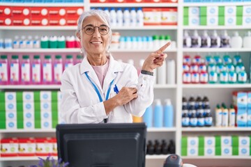 Middle age woman with tattoos working at pharmacy drugstore with a big smile on face, pointing with hand finger to the side looking at the camera.