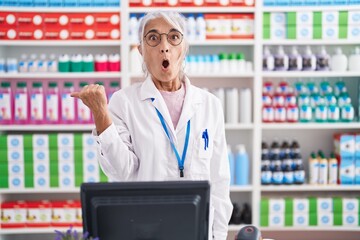 Middle age woman with tattoos working at pharmacy drugstore surprised pointing with hand finger to the side, open mouth amazed expression.