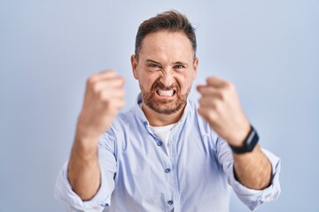 Middle age caucasian man standing over blue background angry and mad raising fists frustrated and furious while shouting with anger. rage and aggressive concept.