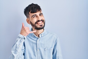 Young hispanic man with beard standing over blue background smiling doing phone gesture with hand and fingers like talking on the telephone. communicating concepts.