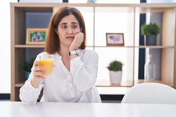 Brunette woman drinking glass of orange juice looking stressed and nervous with hands on mouth biting nails. anxiety problem.