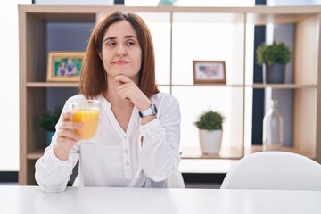 Brunette woman drinking glass of orange juice with hand on chin thinking about question, pensive expression. smiling and thoughtful face. doubt concept.