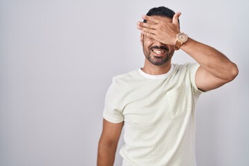 Hispanic man with beard standing over isolated background smiling and laughing with hand on face...