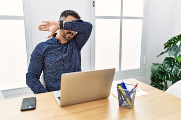 Young hispanic man with beard working at the office with laptop covering eyes with arm, looking...