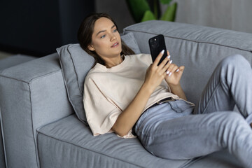 Young woman lying relaxed with phone and laptop on the comfortable couch at home. Working or leisure time at home