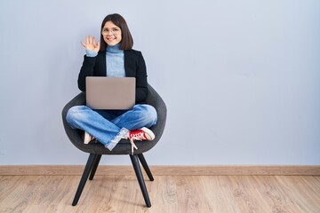 Young hispanic woman sitting on chair using computer laptop waiving saying hello happy and smiling, friendly welcome gesture