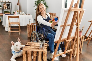 Young caucasian woman smiling confident sitting on wheelchair drawing with dog at art studio