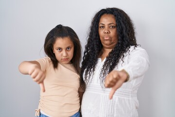 Mother and young daughter standing over white background looking unhappy and angry showing...