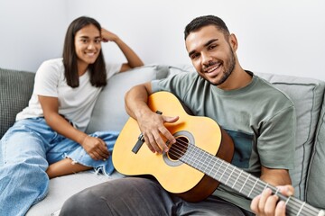 Young latin couple playing classical guitar sitting on the sofa at home.