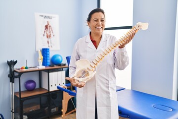 Middle age hispanic woman holding anatomical model of spinal column winking looking at the camera...