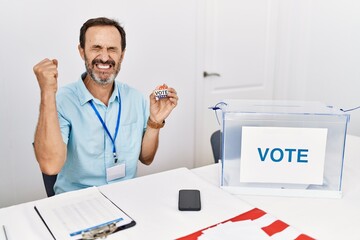 Middle age man with beard sitting by ballot holding i vote badge very happy and excited doing winner gesture with arms raised, smiling and screaming for success. celebration concept.