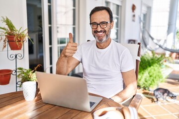 Middle age man using computer laptop at home doing happy thumbs up gesture with hand. approving...