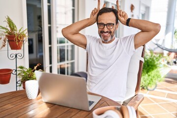 Middle age man using computer laptop at home posing funny and crazy with fingers on head as bunny...
