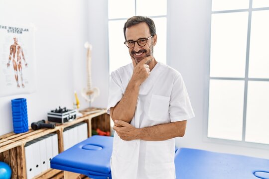 Middle age man with beard working at pain recovery clinic looking confident at the camera smiling with crossed arms and hand raised on chin. thinking positive.