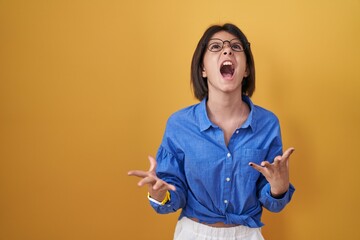 Young girl standing over yellow background crazy and mad shouting and yelling with aggressive...