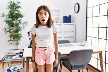 Young hispanic girl standing at pediatrician clinic afraid and shocked with surprise and amazed expression, fear and excited face.