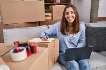 Young woman using laptop write on notebook sitting on sofa at new home