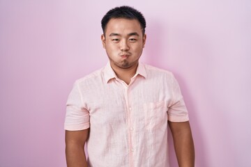 Chinese young man standing over pink background puffing cheeks with funny face. mouth inflated with air, crazy expression.