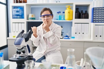 Hispanic girl with down syndrome working at scientist laboratory asking to be quiet with finger on lips pointing with hand to the side. silence and secret concept.