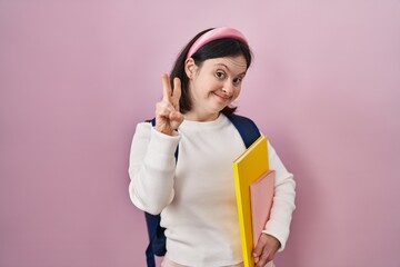 Woman with down syndrome wearing student backpack and holding books smiling with happy face winking at the camera doing victory sign with fingers. number two.