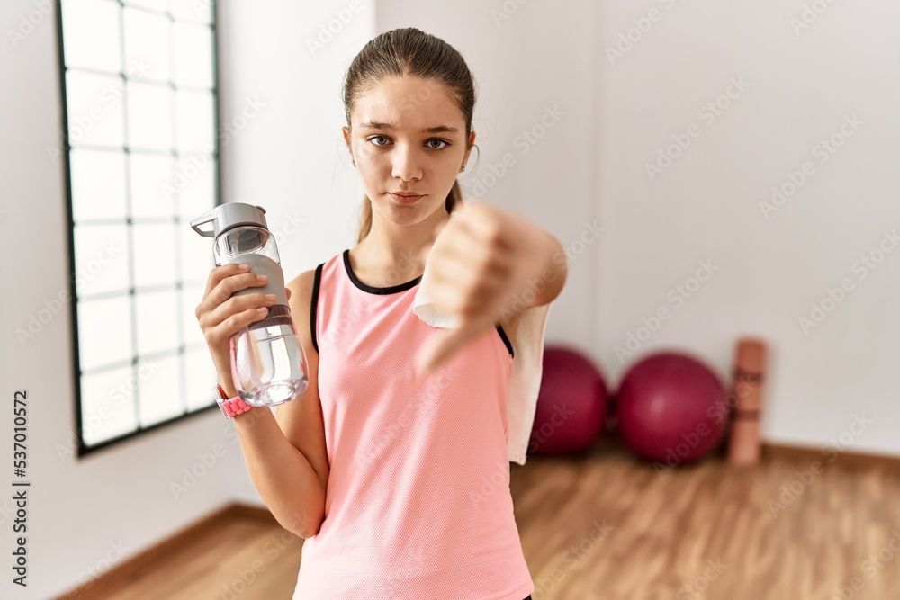 Poster Young brunette teenager wearing sportswear holding water bottle looking unhappy and angry showing rejection and negative with thumbs down gesture. bad expression.