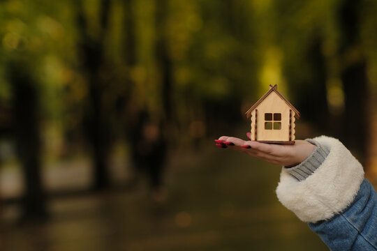 Unrecognizable Woman Holding Small Wooden House In Forest Park. Body Part Of Female With Toy House. Concept Of Purchasing New Apartment, Roof Overhead In Ecological Place.