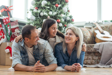 Christmas. Family. Happiness. Top view of dad, mom and daughter in Santa hats looking at camera and smiling while lying on the floor at home.
