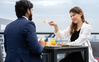 Young couple enjoy breakfast table outdoor on rooftop skyscraper building in cityscape background during sunlight in the morning. Diverse ethnic couple sitting together on roof balcony in skyline view