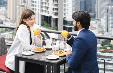 Young couple enjoy breakfast table outdoor on rooftop skyscraper building in cityscape background during sunlight in the morning. Diverse ethnic couple sitting together on roof balcony in skyline view
