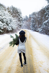 A girl in a dress and hat stands with her back in the middle of a winter road in the forest.