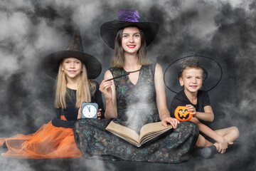 A happy family in creepy costumes of witches and wizards, holding large book with predictions, baskets in the shape of a pumpkin, a black watch and a magic wand on a black smoky background