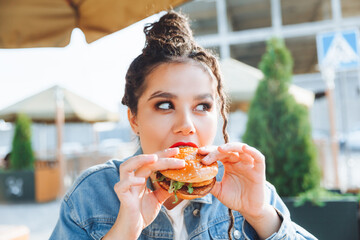 A young glamorous woman with dreadlocks and red lipstick is sitting and eating a burger in a street cafe, the concept of eating. long-lasting lipstick