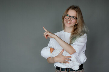 Portrait of young cheerful woman holding baby doll in white clothes, pointing with index finger on...