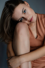 Young flawless blue-eyed woman in beige bodysuit sitting on floor, hugging knees on grey background, looking at camera.