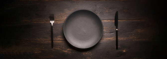 A black empty plate and a fork with a knife on a dark background. kitchen restaurant invitation...