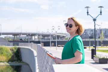 A smiling middle-aged woman in sunglasses with a notebook or tablet in her hands on city embankment. Break from work.A woman walks around the city during her vacation. The concept of urban tourism