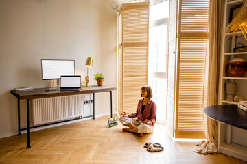 Young woman meditates after work, sitting with tablet on the floor at cozy home office. Stylish interior with workplace and computers. Mental health and relaxation, working from home concept