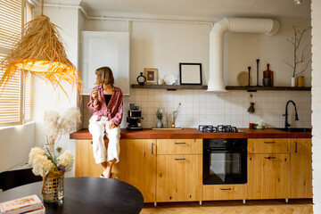 Stylish kitchen interior of modern apartment made in white and beige tones with wooden kitchen front and floor. Woman sitting with coffee on kitchen table top © rh2010