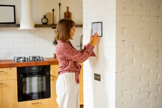 Woman controls room temperature on a digital panel mounted on the wall in kitchen. Concept of smart home and new technologies for home comfort