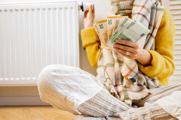 Warmly dressed woman counting money while sitting near radiator at home. Concept of expensive energy resources and the energy crisis in Europe