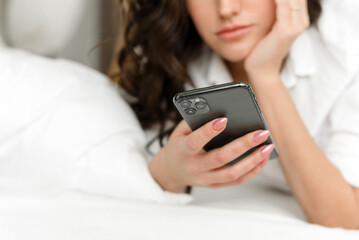 A young woman is checking social networks and news on her phone while lying on the bed. The woman...