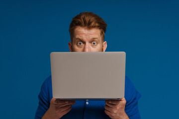 Ginger shocked man looking at camera while posing with laptop