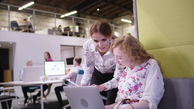 Woman working in creative office, using laptop, colleague helping