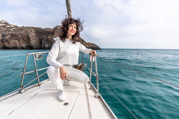 A woman sits on the bow of a yacht on a sunny summer day, the breeze develops her hair, a beautiful sea is in the background