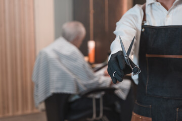 Detalle de peluquero irreconocible, enseñando las tijeras y de fondo cliente senior, leyendo una revista a la espera de cortarse el pelo.