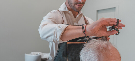 Hombre peluquero irreconocible, cortando el pelo con tijera, a cliente senior anónimo, en la peluquería. Fotografía horizontal con espacio para texto. - obrazy, fototapety, plakaty