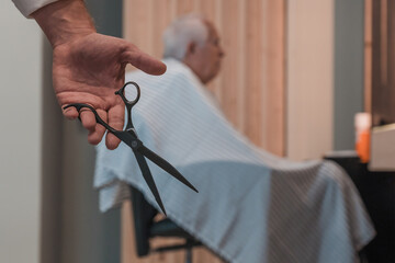 Detalle de mano de peluquero con tijeras, y de segundo plano, cliente senior, sentado para cortar el pelo. - obrazy, fototapety, plakaty