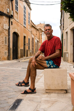 Older Traveling Man Sitting Looking At The Camera In A Picturesque Village In Spain.