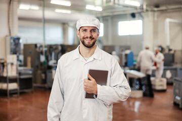 A happy food factory supervisor holds tablet in hands and smiles at the camera. He is on his way to assess the quality of the food.