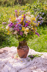 Lush multi-colored bouquet of wild flowers in a jug on a wooden table against the background of greenery in a rustic style